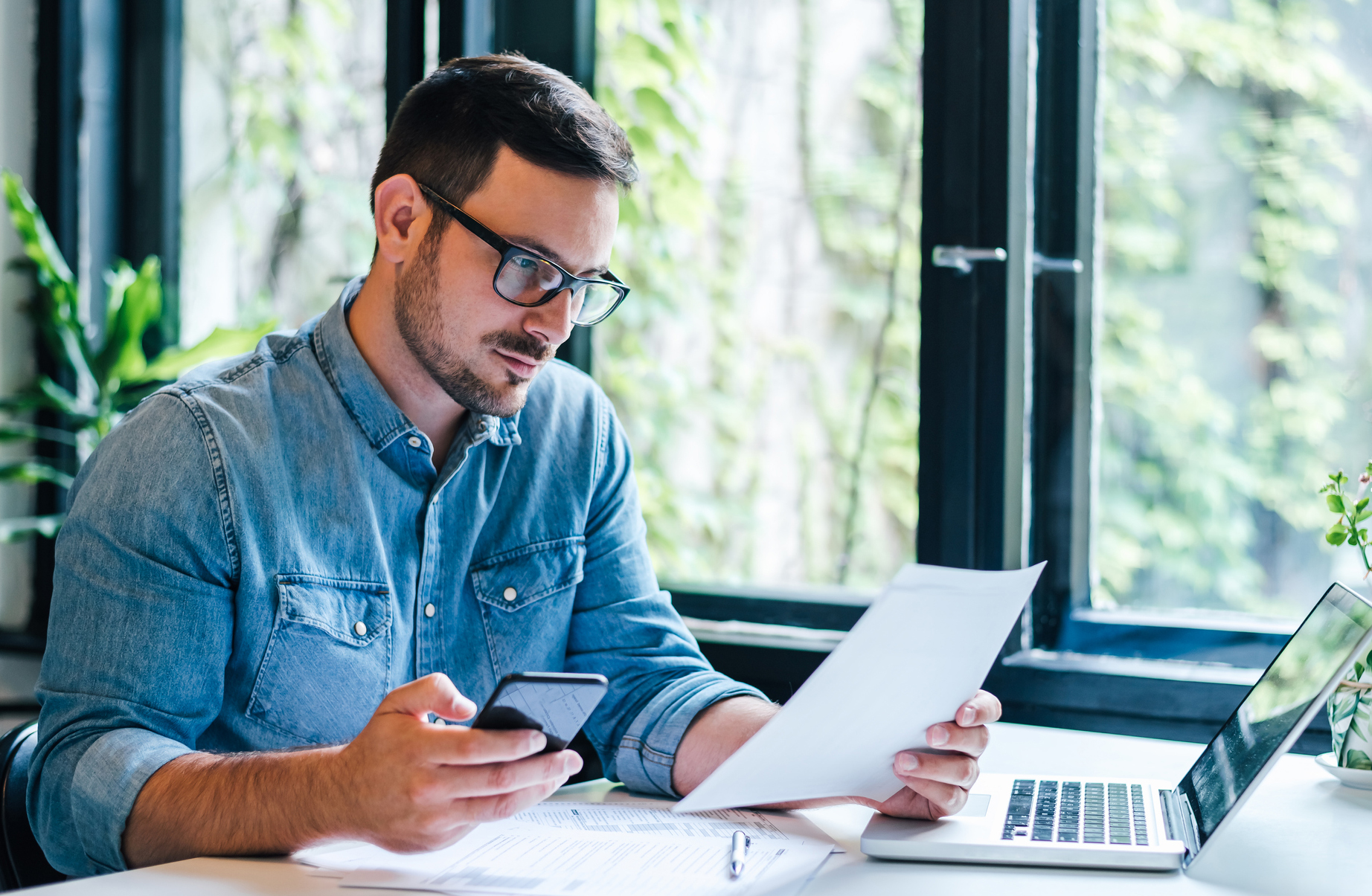 Serious pensive thoughtful focused young casual entrepreneur small business owner accountant bookkeeper in office looking at and working with laptop and income tax return papers and documents